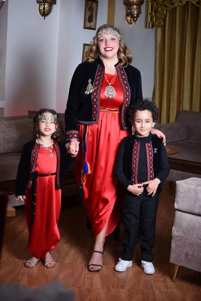 Femme et deux enfants en tenues traditionnelles algériennes rouges avec vestes noires brodées dans un salon.